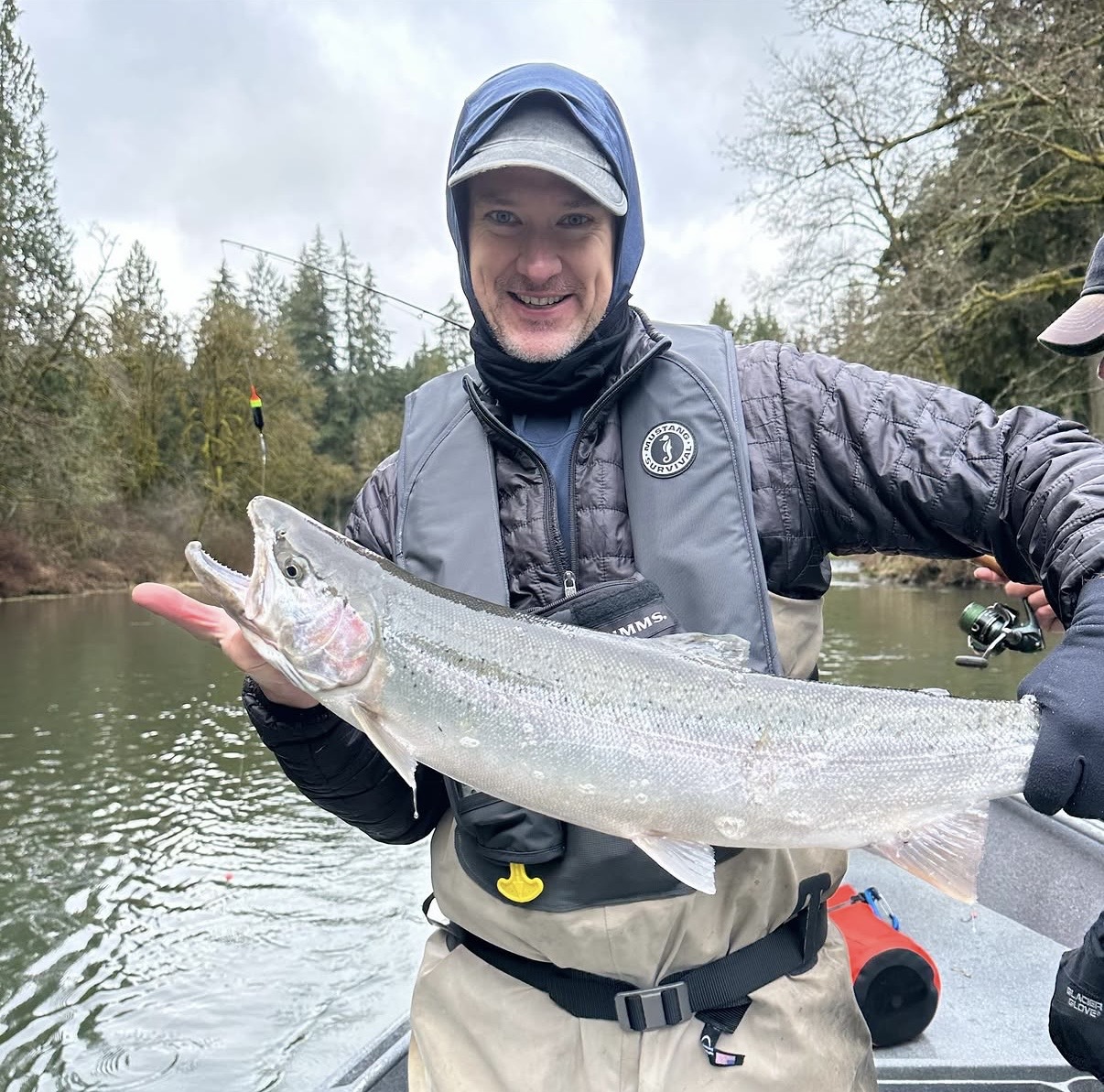 Angler in rain gear holding a chrome-bright steelhead aboard the boat on a misty winter river